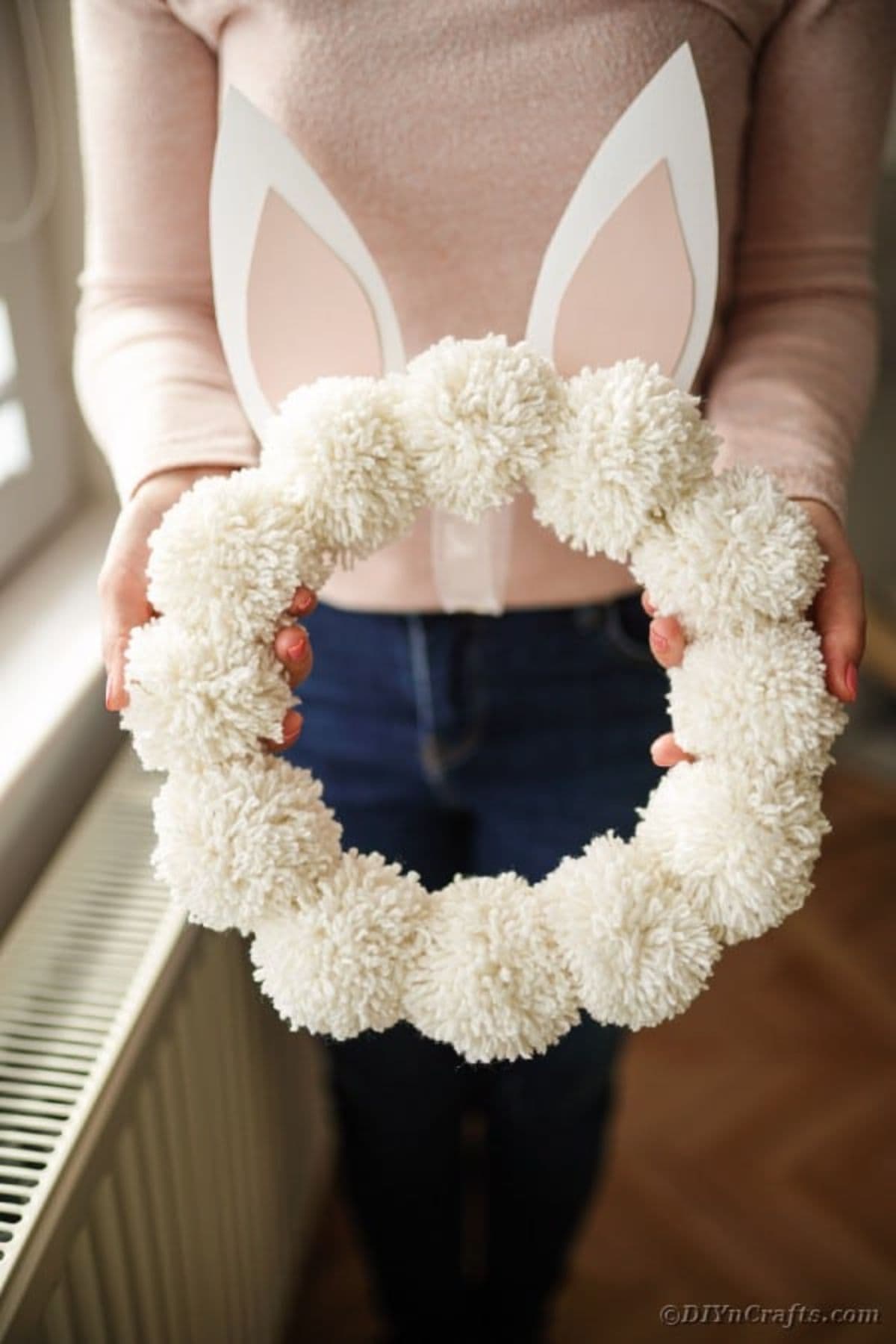 a woman in a white top and blue jeans is holding a wreath made of white pom poms with white bunny ears on the top