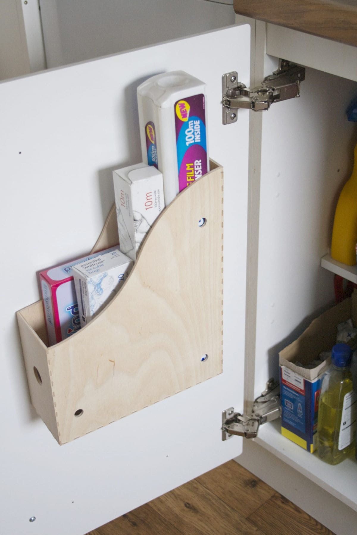 The inside of a kitchen cabinet is shown. On the door a wooden magazine rack has been fixed and rolls of cling film and foil put inside